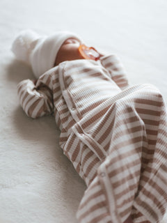 A baby dressed in a striped outfit and white hat, resting on a soft surface with a pacifier.