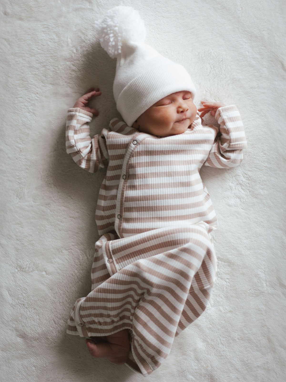 Newborn baby in a striped outfit and fluffy white hat, sleeping peacefully on a soft surface.