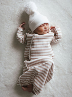 Newborn baby in a striped outfit and fluffy white hat, sleeping peacefully on a soft surface.