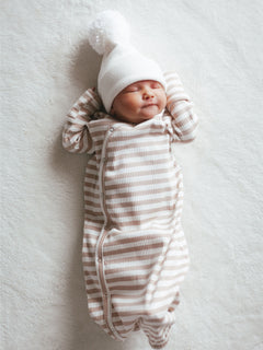 Smiling newborn in a striped onesie and pom-pom hat, laying on a soft blanket.