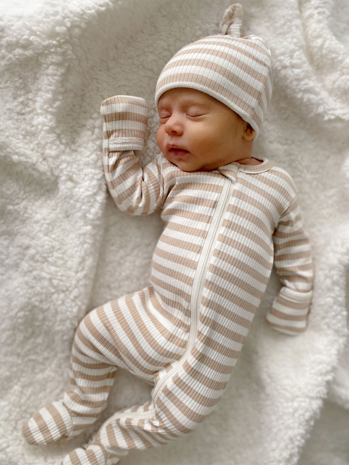 Newborn in striped onesie and matching hat, sleeping peacefully on a soft white blanket.