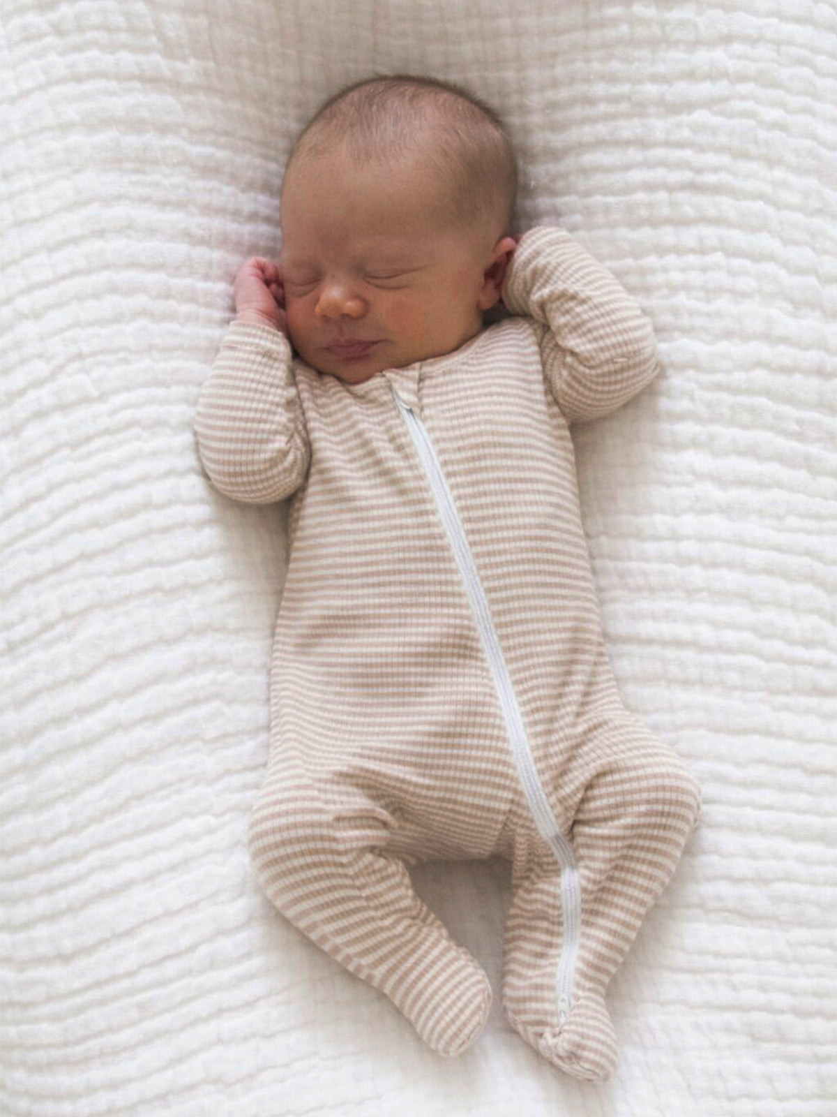 Newborn baby sleeping peacefully on a soft, textured blanket, wearing a striped zip-up onesie.
