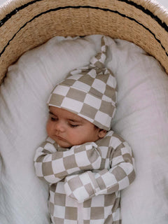 Sleeping baby in a checkered outfit and hat, nestled in a woven bassinet on soft blankets.