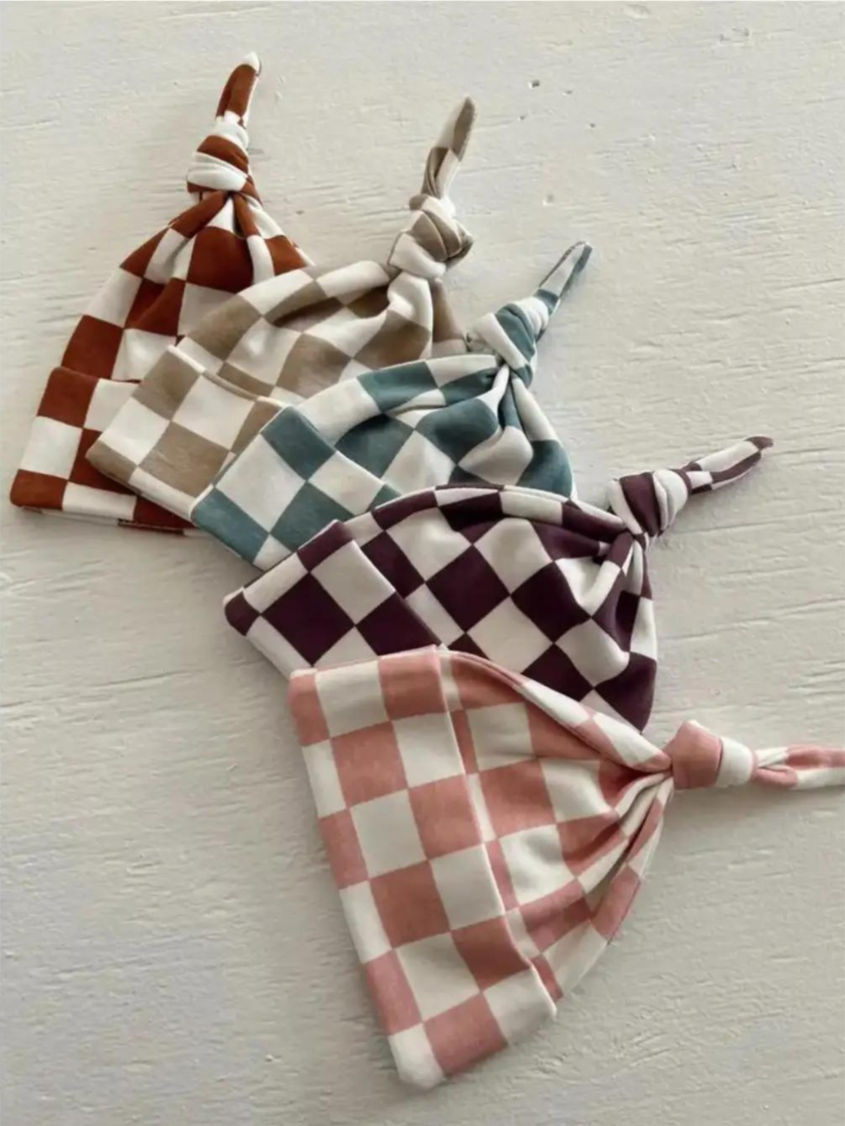 Four checkered baby hats in earthy tones laid on a textured white surface.