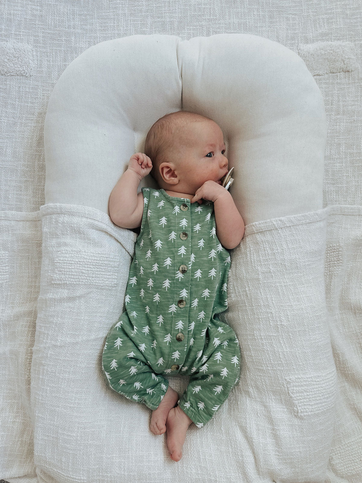 Baby in green tree-patterned outfit lying on a soft, white blanket.