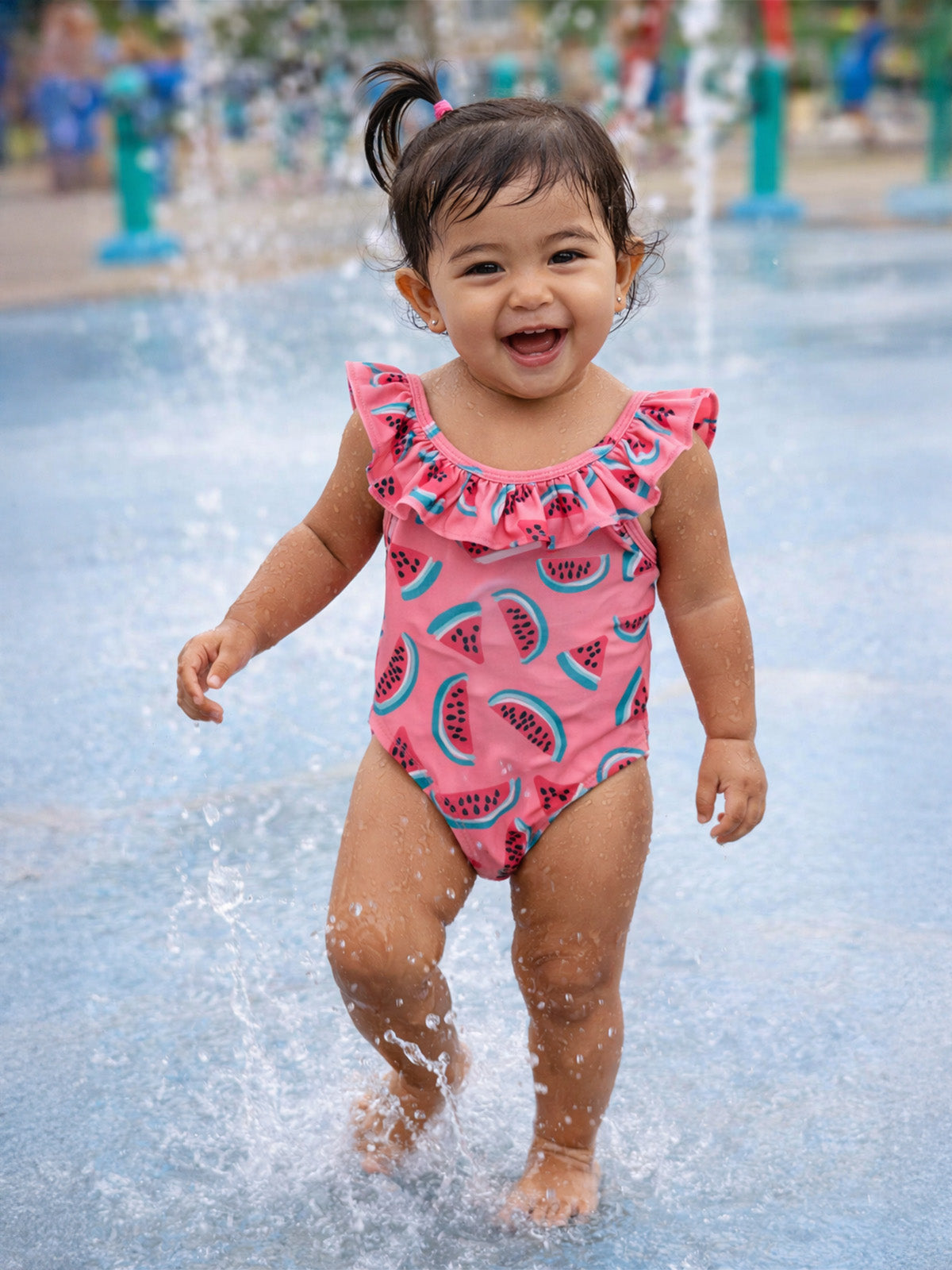 Smiling toddler in a watermelon-patterned swimsuit playing in a splash pad.